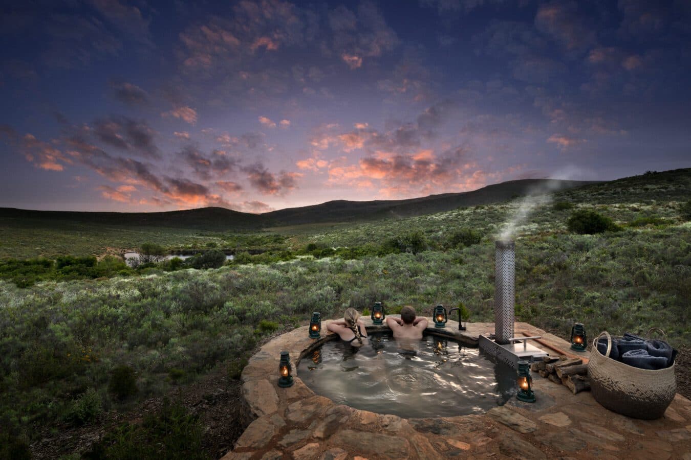 Melozhori Main Lodge: Couple in steaming stone hot tub, pink dusk over karoo valley